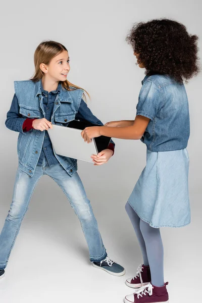Excited kid in stylish denim clothes taking laptop away from african american friend on grey — Stock Photo