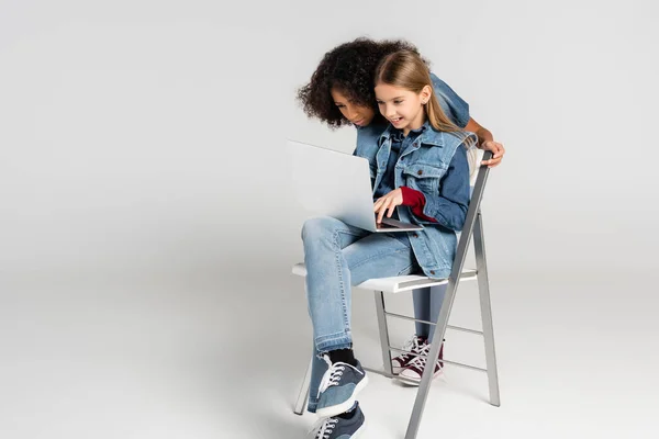 Fröhliches Kind in modischen Jeansklamotten auf Stuhl sitzend und mit Laptop in der Nähe eines afrikanisch-amerikanischen Freundes auf grau — Stockfoto