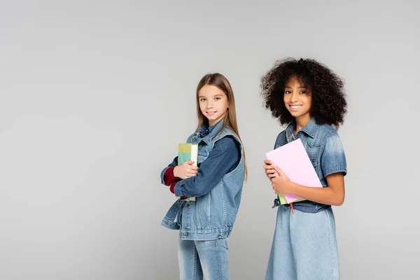 Zwei fröhliche multikulturelle Schulkinder in Jeanskleidung stehen mit Büchern isoliert auf grau — Stockfoto