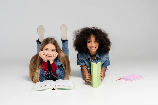 Cheerful schoolkids smiling at camera while lying with textbooks on grey — Stock Photo