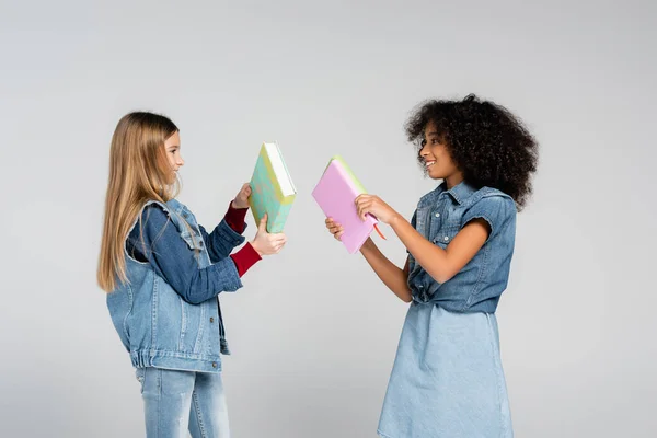 Side view of happy schoolgirls in denim clothes looking at each other while holding books isolated on grey — Stock Photo