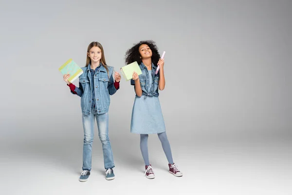 Full length view of fashionable multicultural schoolgirls standing with books on grey — Stock Photo