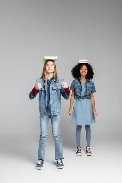 Smiling interracial schoolgirls in denim clothes and gumshoes standing with books on heads on grey — Stock Photo