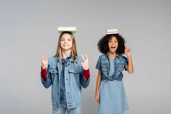 Emocionado colegiala afroamericana señalando con el dedo cerca amigo feliz mostrando signos de victoria aislado en gris - foto de stock