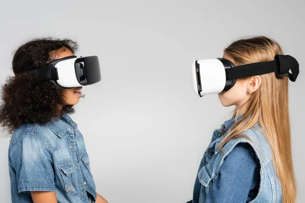 Side view of fashionable interracial kids in vr headsets looking at each other isolated on grey — Stock Photo