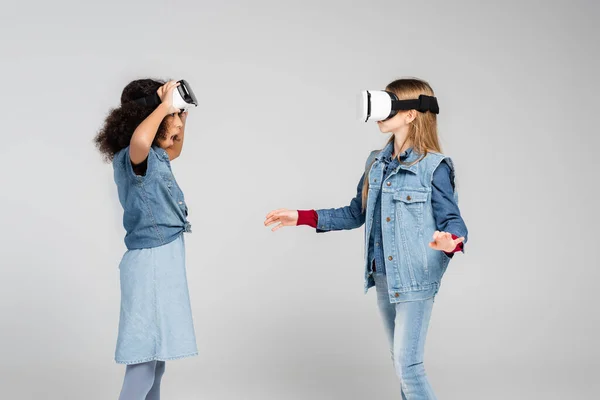 Amazed african american girl looking at friend gaming in vr headset on grey — Stock Photo