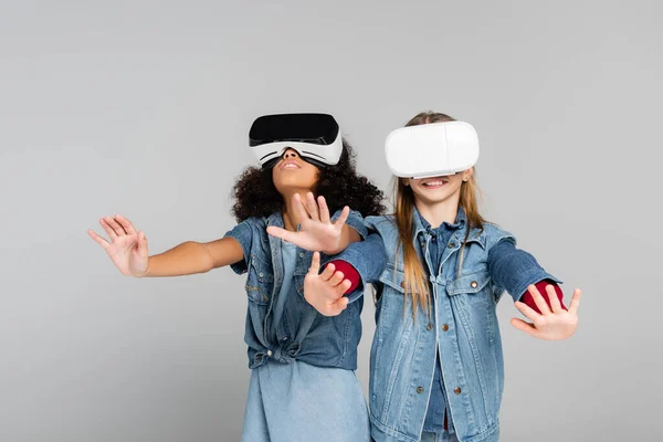 Excited multicultural kids in stylish denim clothes gesturing in vr headsets on grey — Stock Photo