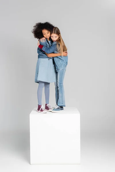 Astonished interracial girls in trendy denim clothes embracing while standing on white cube on grey — Stock Photo