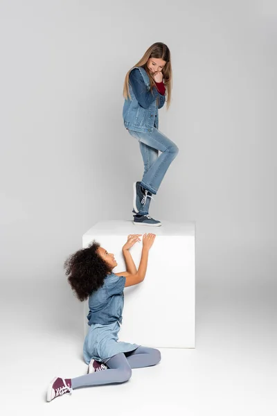 Cheerful girl standing on white cube while african american girl sitting on grey — Stock Photo
