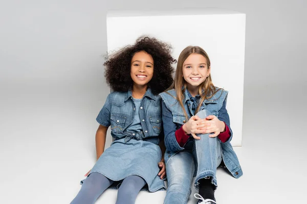 Trendy interracial girls smiling at camera while sitting near white box on grey — Stock Photo