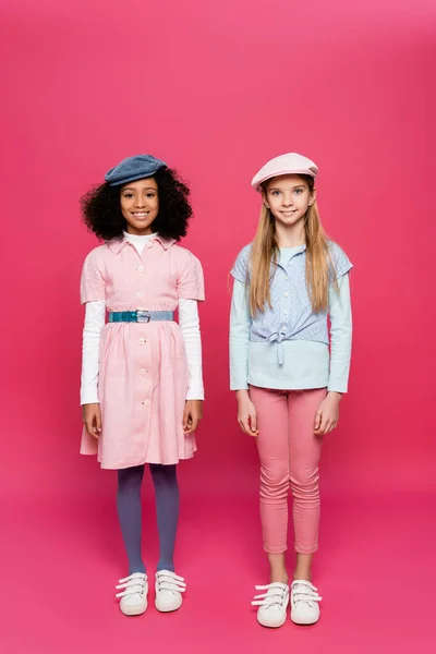 Visão de comprimento total de duas meninas inter-raciais felizes em roupas da moda posando em rosa — Fotografia de Stock