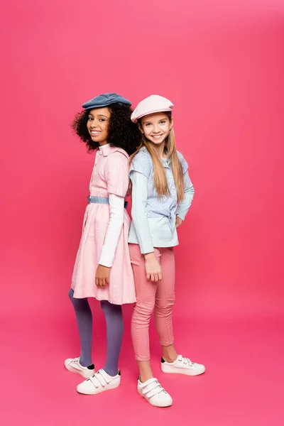 Visão de comprimento total de meninas multiculturais alegres em roupas na moda e bonés olhando para a câmera no rosa — Fotografia de Stock