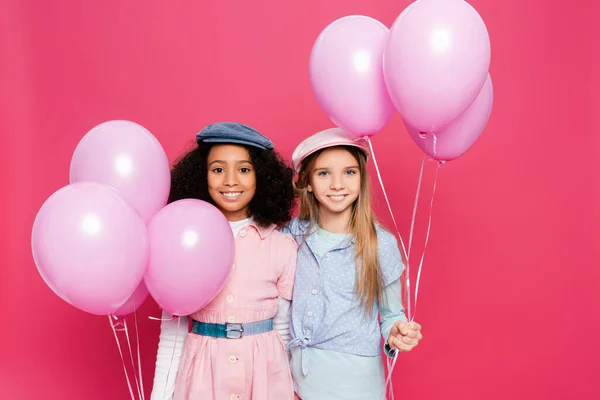Duas meninas multiculturais felizes em roupa de primavera na moda posando com balões festivos isolados em rosa — Fotografia de Stock