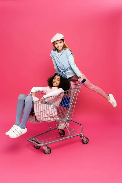 Fröhliche gemischtrassige Mädchen im trendigen Outfit haben Spaß mit Einkaufswagen auf rosa — Stockfoto