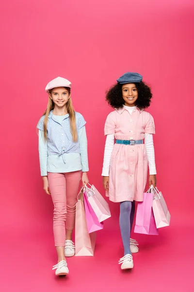 Stilvolle multikulturelle Mädchen lächeln in die Kamera, während sie mit Einkaufstaschen auf rosa laufen — Stockfoto
