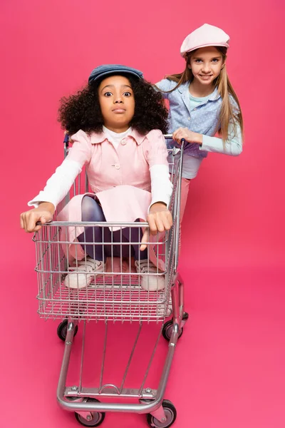 Erstaunt afrikanisch-amerikanisches Kind sitzt im Einkaufswagen neben glücklichen Freund auf rosa — Stockfoto