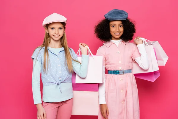 Fröhliche interrassische Kinder in modischen Klamotten posieren mit Einkaufstaschen auf rosa — Stockfoto