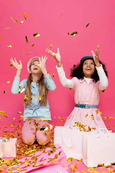 Asombrado, chicas interracial de moda lanzando confeti cerca de regalos en rosa - foto de stock