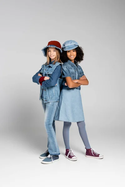 Stylish interracial girls in denim clothes and gumshoes standing with crossed arms on grey — Stock Photo