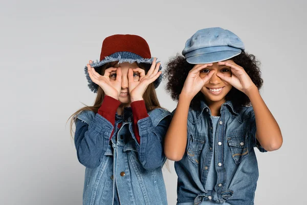 Joyful multicultural friends in denim clothes and hats showing eyeglasses with hands isolated on grey — Stock Photo