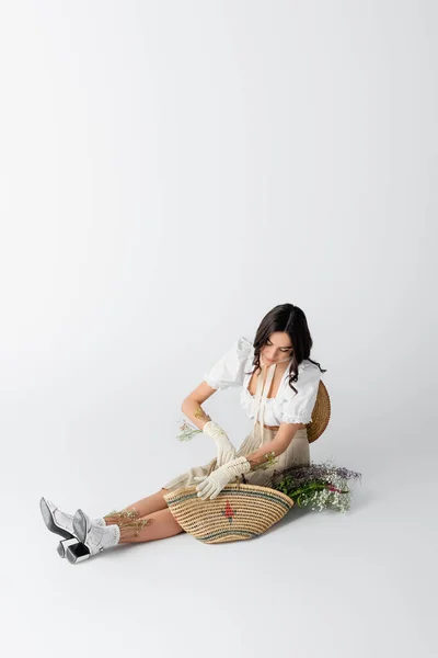 Young woman in spring outfit and gloves sitting near flowers in straw bag on white — Stock Photo