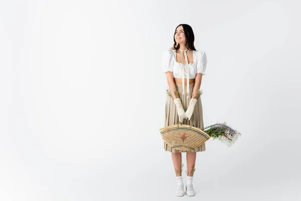 Piena lunghezza di gioiosa giovane donna in abito primavera che tiene borsa di paglia con fiori su bianco — Foto stock