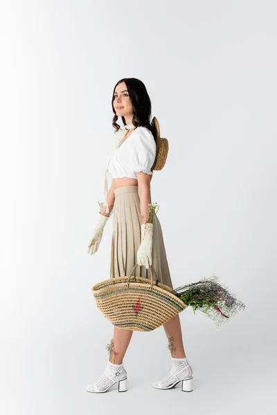 Full length of happy woman in spring outfit holding straw bag with flowers on white — Stock Photo