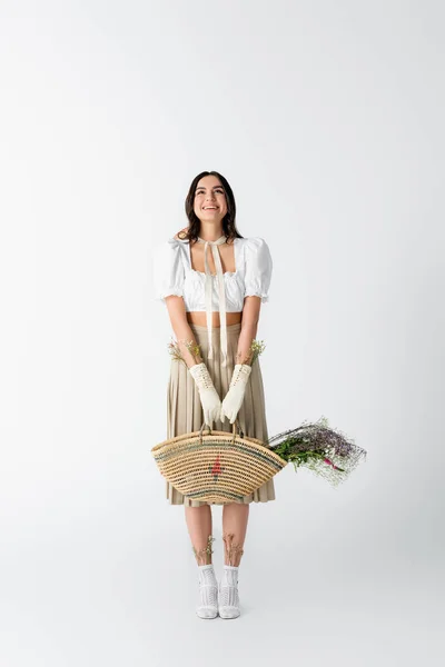 Piena lunghezza di felice giovane donna in abito primavera in possesso di borsa di paglia con fiori su bianco — Foto stock