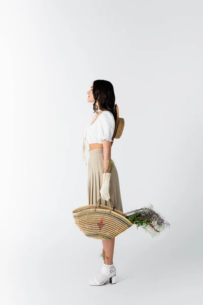 Vue latérale de la jeune femme en tenue de printemps tenant sac de paille avec des fleurs sur blanc — Photo de stock