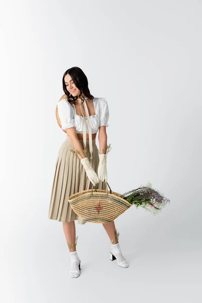 Piena lunghezza di donna felice in abito primaverile sorridente mentre tiene borsa di paglia con fiori su bianco — Foto stock