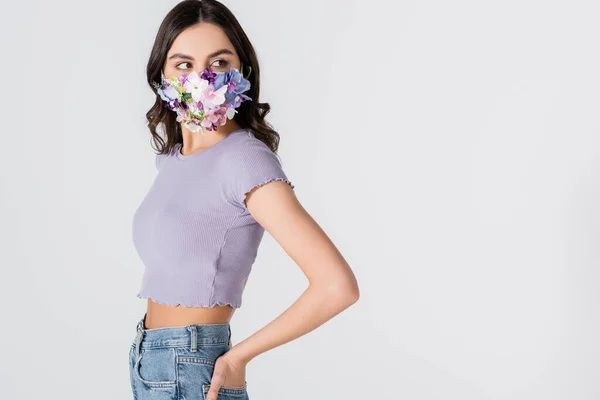 Junge Frau in bauchfreiem Top und medizinischer Maske mit blühenden Blumen, die vereinzelt auf weißem Grund wegschauen — Stockfoto