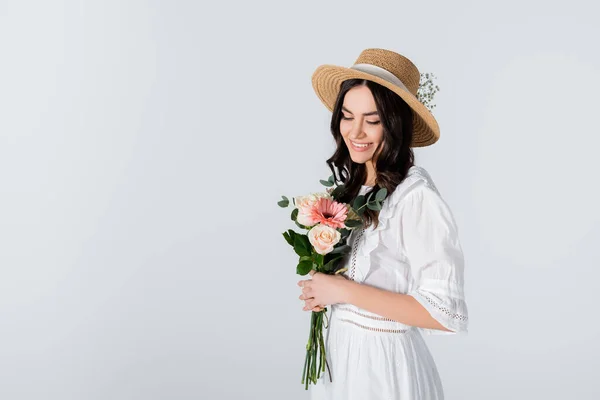 Cheerful young woman in straw hat and dress holding bouquet of spring flowers isolated on white — Stock Photo