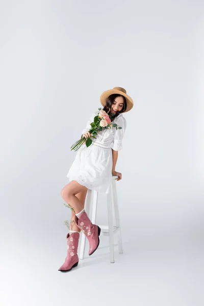 Gioiosa donna in cappello di paglia e vestito seduto sulla sedia e tenendo bouquet di fiori primaverili su bianco — Foto stock