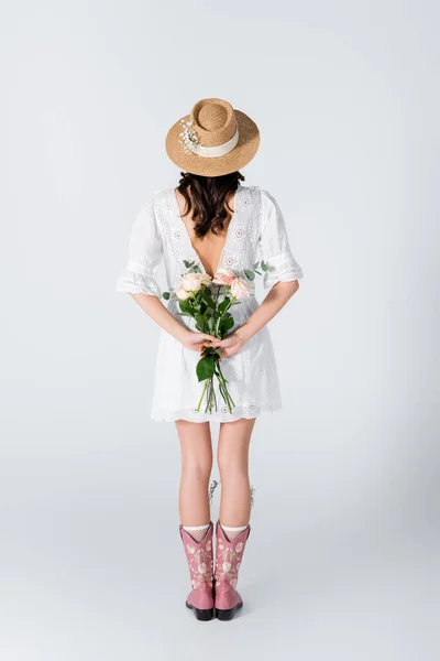 Vista posteriore della giovane donna in cappello di paglia e vestito che tiene bouquet di fiori primaverili su bianco — Foto stock