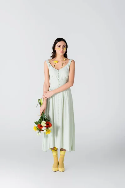 Full length of brunette woman with petals on face holding bouquet of flowers on white — Stock Photo