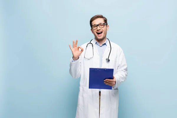 Verblüffter junger Arzt mit Brille und weißem Mantel hält Klemmbrett und zeigt Okay-Zeichen auf blau — Stockfoto