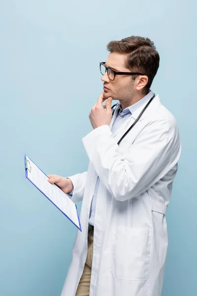 Nachdenklicher junger Arzt mit Brille und weißem Mantel, Klemmbrett isoliert auf blauem Grund — Stockfoto