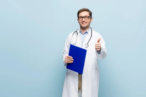 Médico alegre en gafas y abrigo blanco apuntando con el dedo al portapapeles y mostrando el pulgar hacia arriba en azul - foto de stock