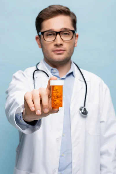 Blurred doctor in glasses and white coat holding bottle with medication isolated on blue — Stock Photo