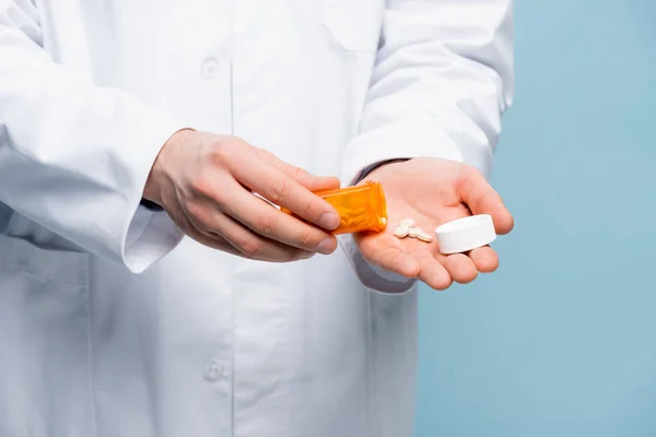 Cropped view of doctor in white coat holding pills and bottle in hands isolated on blue — Stock Photo