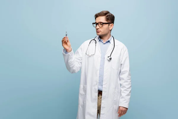 Médico joven en gafas y abrigo blanco mirando la jeringa en azul - foto de stock