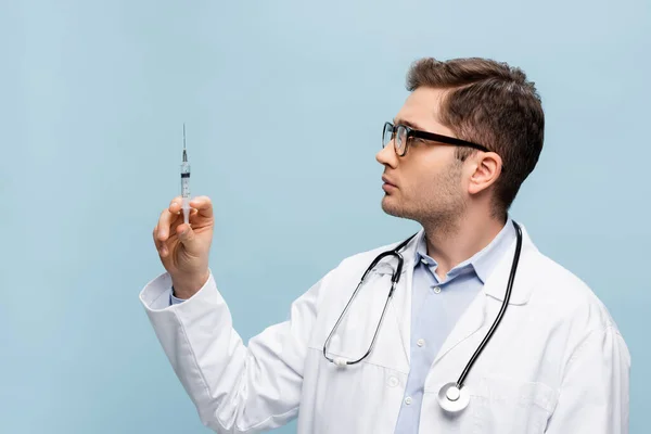 Médico en gafas y abrigo blanco mirando la jeringa aislada en azul - foto de stock