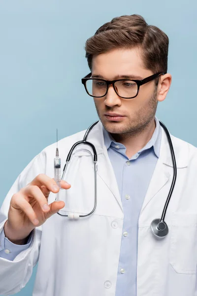 Doctor in glasses and white coat looking at syringe with vaccine isolated on blue — Stock Photo