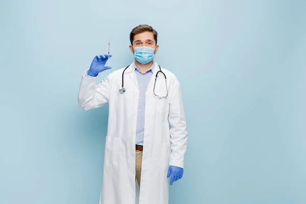 Young doctor in medical mask and white coat with stethoscope holding syringe on blue — Stock Photo