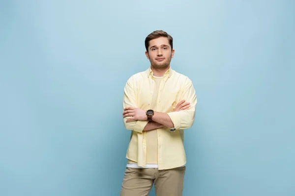 Young man in yellow shirt looking at camera and standing with crossed arms isolated on blue — Stock Photo