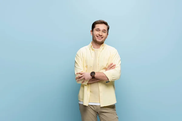 Glücklicher junger Mann im gelben Hemd, der in die Kamera blickt und mit verschränkten Armen auf blau steht — Stockfoto