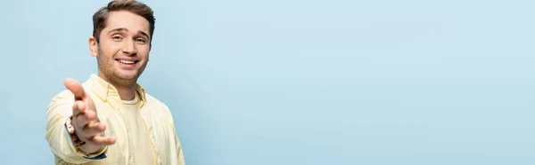 Cheerful young man in yellow shirt with blurred outstretched hand isolated on blue, banner — Stock Photo