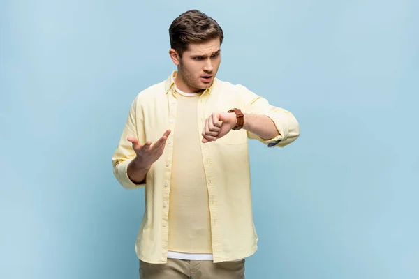 Mécontent jeune homme en chemise jaune regardant montre-bracelet sur bleu — Photo de stock