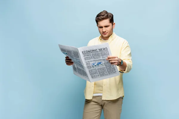 Displeased young man in shirt reading economical newspaper on blue — Stock Photo