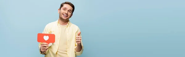 Homem feliz na camisa segurando coração de papel vermelho e mostrando polegar para cima isolado em azul, banner — Fotografia de Stock